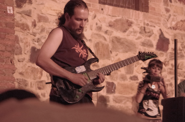 Ein bärtiger Mann spielt eine Gitarre auf der Bühne, rechts von ihm hält eine Frau ein Mikrofon und im Hintergrund ist eine Menschenmenge zu sehen.