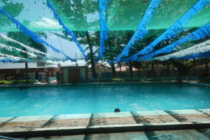 Großer Swimmingpool mit blauen und grünen Bändern, die von der Decke hängen, umgeben von Häusern, Bäumen und einem klaren blauen Himmel, mit Menschen im Wasser und in der Sonne.