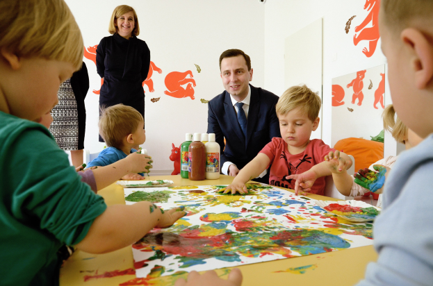 Kinder sitzen um einen Tisch mit Farbe an den Händen, beschäftigt mit einer kreativen Aktivität mit Papieren und Flaschen auf dem Tisch, während eine Frau im Hintergrund lächelt mit Gemälden an der Wand.