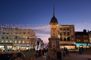 Eine belebte Stadtstraße bei Nacht mit Menschen, Fahrzeugen, Fahrrädern, Mülltonnen, Gebäuden, Laternen, Ampeln und einem Uhrenturm im Hintergrund, beleuchtet von festlichen Weihnachtslichtern.