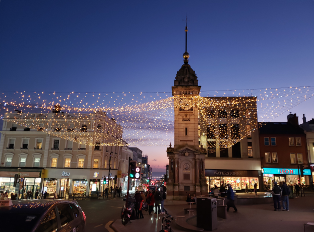 Eine belebte Stadtstraße bei Nacht mit Menschen, Fahrzeugen, Fahrrädern, Mülltonnen, Gebäuden, Laternen, Ampeln und einem Uhrenturm im Hintergrund, beleuchtet von festlichen Weihnachtslichtern.