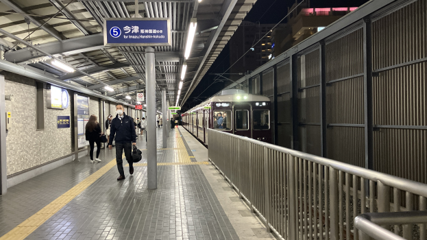 Ein Zug fährt auf ein Bahnsteig, wo Menschen gehen, mit einem Geländer auf der rechten Seite und Gebäuden im Hintergrund; überkopfleuchten und eine Abfahrtsanzeige sind auf dem Bahnsteig zu sehen.