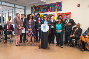 Gruppe von Menschen vor einem Podium mit Mikrofon und einer Tafel mit Text, einige sitzen auf Stühlen und andere stehen, mit Flaggen, einer Tafel, einem Feuerlöscher und Deckenleuchten an der Rückwand.