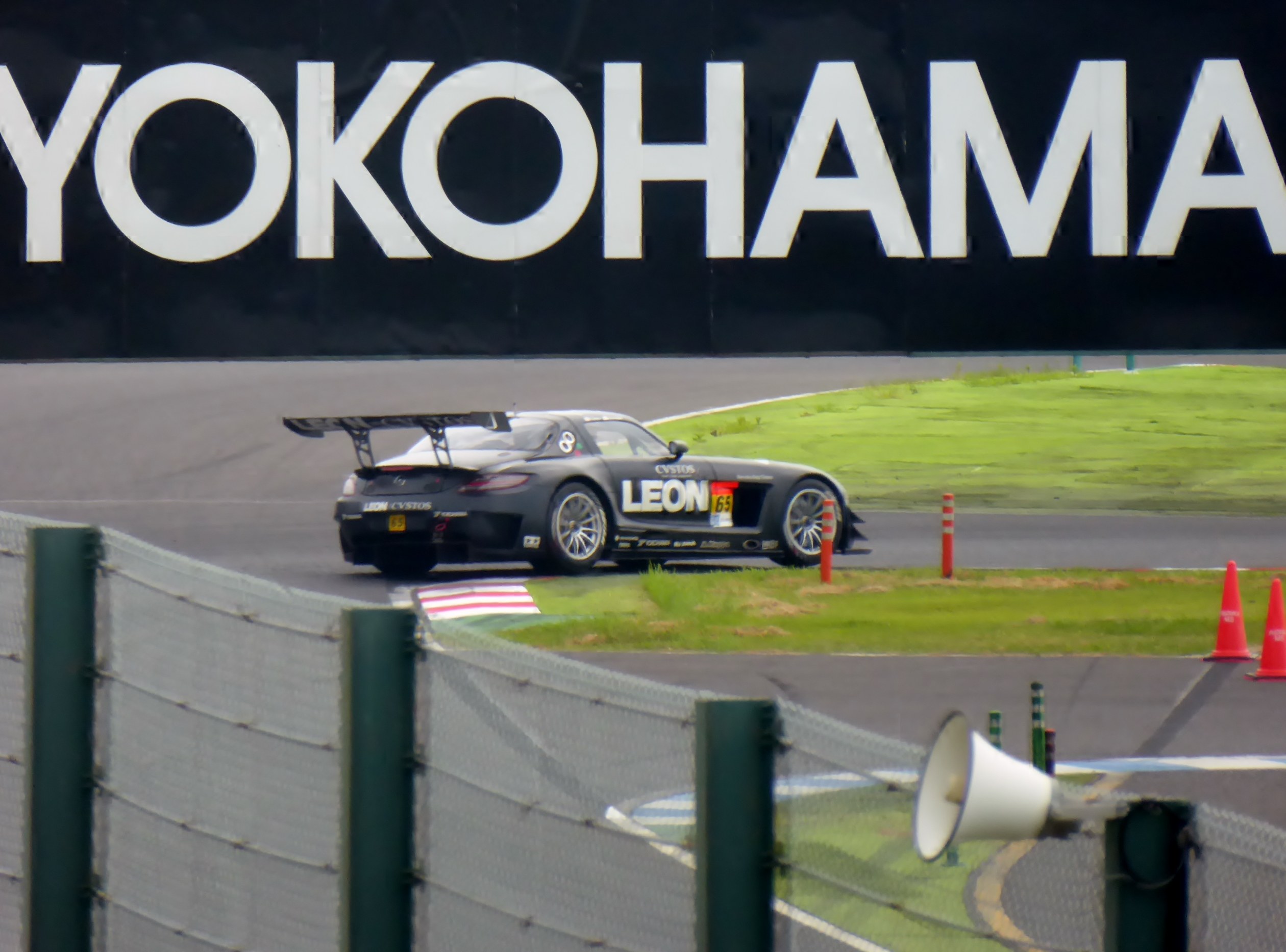 Schwarzer Sportwagen auf einer Rennstrecke mit einem "Yokohama"-Schild im Hintergrund, umgeben von Verkehrskegeln, Gras und einem Zaun, mit einem Lautsprecher im Vordergrund.