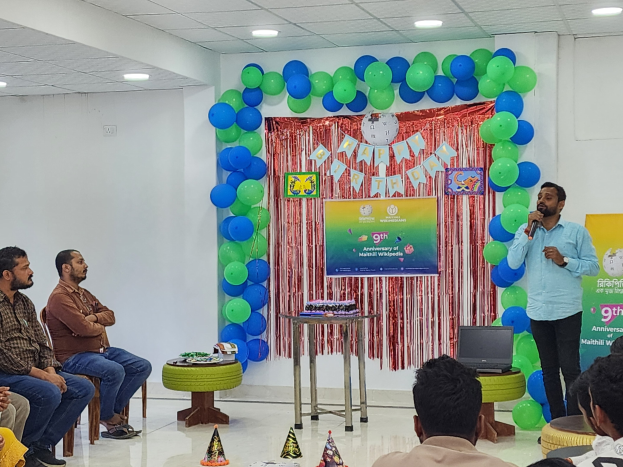 Eine Gruppe von Menschen sitzt auf Stühlen und schaut auf eine Bühne mit Ballons, einem Banner, einer Uhr und anderen Gegenständen, während eine Person auf der Bühne ein Mikrofon in der Nähe eines Tisches mit einem Laptop und anderen Gegenständen hält, unter Deckenlampen während einer 9. Geburtstagsfeier.