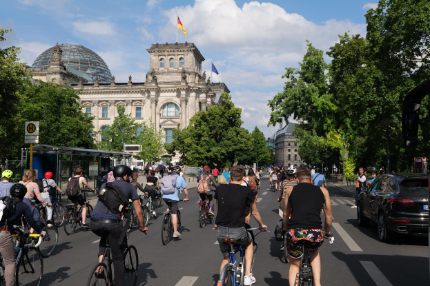Eine Gruppe von Menschen, die auf Fahrrädern eine von Bäumen gesäumte Straße in Berlin, Deutschland, entlangfahren, mit Gebäuden und einer Bushaltestelle im Hintergrund, unter einem bewölkten Himmel und einer Flagge an einem Gebäude.