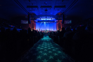 Ein gro├če Auditorium mit sitzenden Zuschauern, eine Bühne mit Musikern und überkopfleuchten und Fernseher in einer dunklen Konzerthallenatmosph├Ąre, wahrscheinlich für die Nationale Convention 2019.
