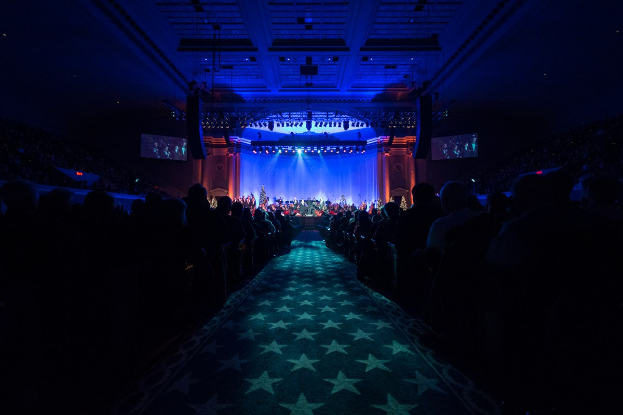 Ein gro├če Auditorium mit sitzenden Zuschauern, eine Bühne mit Musikern und überkopfleuchten und Fernseher in einer dunklen Konzerthallenatmosph├Ąre, wahrscheinlich für die Nationale Convention 2019.