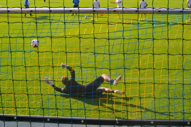 Fußballspieler beim Abwehren eines Schusses mit einem Tor im Vordergrund und einigen Menschen und Bällen im Hintergrund.
