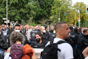 Eine große Gruppe von Menschen, einige mit Masken und Rucksäcken oder Kameras, steht vor einer Gruppe von Polizeibeamten, mit Laternenmasten, Ampeln, Verkehrszeichen, Bäumen und einem klaren blauen Himmel im Hintergrund.