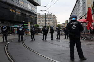 Eine Gruppe von Polizeibeamten in schwarzen Uniformen und Helmen steht auf einer Straße, die von Gebäuden, Bäumen und verstreuten Outdoor-Möbeln gesäumt ist, unter einem klaren Himmel.