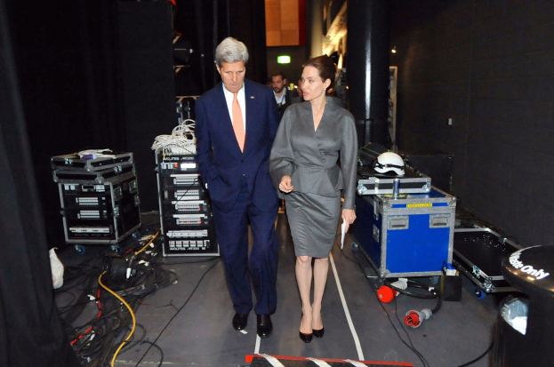 Angelina Jolie und John Kerry auf einem Boden umgeben von einer Gruppe von Menschen, verschiedenen Geräten, Kabeln, einem Helm auf einem Wagen, einem Müllbehälter, einem Vorhang, einer Säule, einem Schild und einer Decke mit Deckenleuchten.