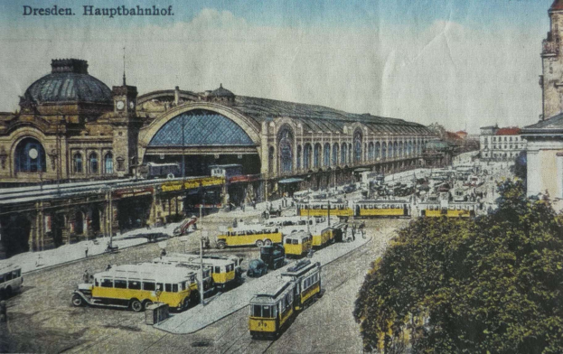Ein sepiafarbenes Postkartenbild des Dresdner Hauptbahnhof in Deutschland, das eine belebte Straße mit Fahrzeugen, Fußgängern, Bäumen, Gebäuden und einem bewölkten Himmel zeigt, mit der Aufschrift "Dresden Hauptbahnhof" oben.