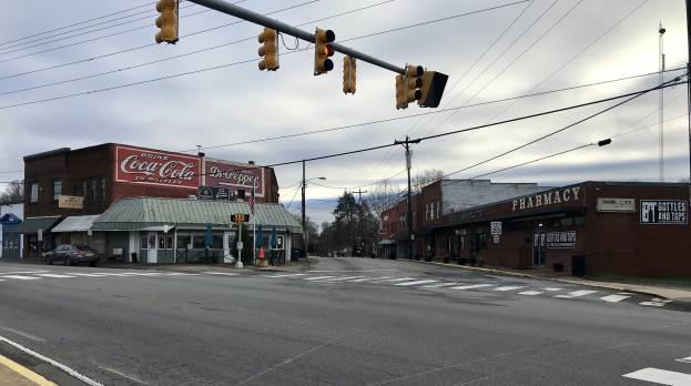 Stadtstraßenkreuzung mit Ampel, Fahrzeugen, Gebäuden mit Schildern, Strommasten mit Drähten, Bäumen und bewölktem Himmel.