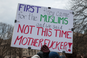 Eine Gruppe von Menschen mit Mützen und Handschuhen hält ein Schild mit der Aufschrift "Zuerst kamen sie wegen der Muslime und wir sagten nicht dieses Mal, verdammte Scheiße", mit Bäumen, Gebäuden und einem klaren blauen Himmel im Hintergrund.