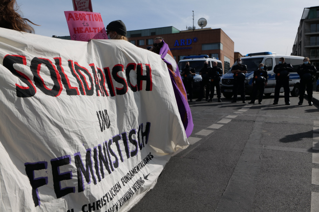 Gruppe von Menschen, die eine "Solidarität und Feminismus"-Schlagzeile tragen, marschieren auf einer Straße mit parkenden Fahrzeugen, Gebäuden, einer Schüsselantenne und einem klaren blauen Himmel im Hintergrund.