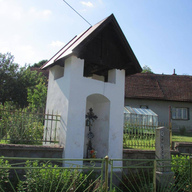 Kleines weißes Kirchengebäude mit einem Kreuz darauf, umgeben von einem Metallzaun, Pflanzen, Gras, einem Weg, nahen Häusern, Polen, Drähten und einem bewölkten Himmel, identifiziert als das Heilig-Geist-Kirchlein.