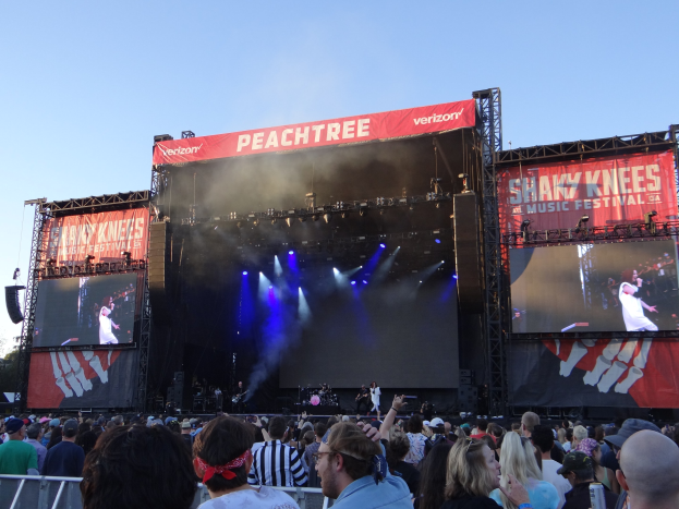 Eine große Menge steht vor einer hell erleuchteten Bühne auf dem Shaky Knees Music Festival, wo Musiker unter Bannern spielen, mit einem Geländer im Vordergrund und einem klaren blauen Himmel im Hintergrund.