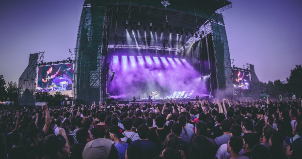 Eine große Menge Menschen bei einem Musikfestival schaut auf einer hell erleuchteten Bühne mit Instrumenten und Bildschirmen, die vor einem Gebäude, Bäumen und einem klaren blauen Himmel liegt.