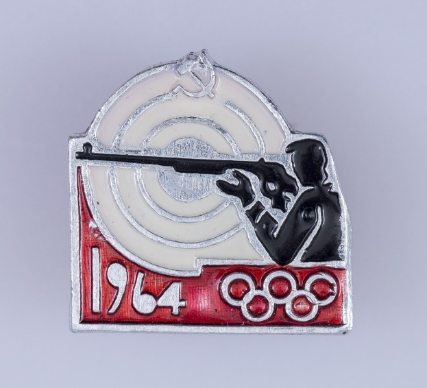 Olympische Spiele-Badge mit einem Mann in rot-weiß gestreiftem Hemd und schwarzem Hut, der ein Gewehr auf ein Ziel richtet, mit dem Text "1964 Olympische Spiele", vor einem hellblauen Himmel mit weißen Wolken.