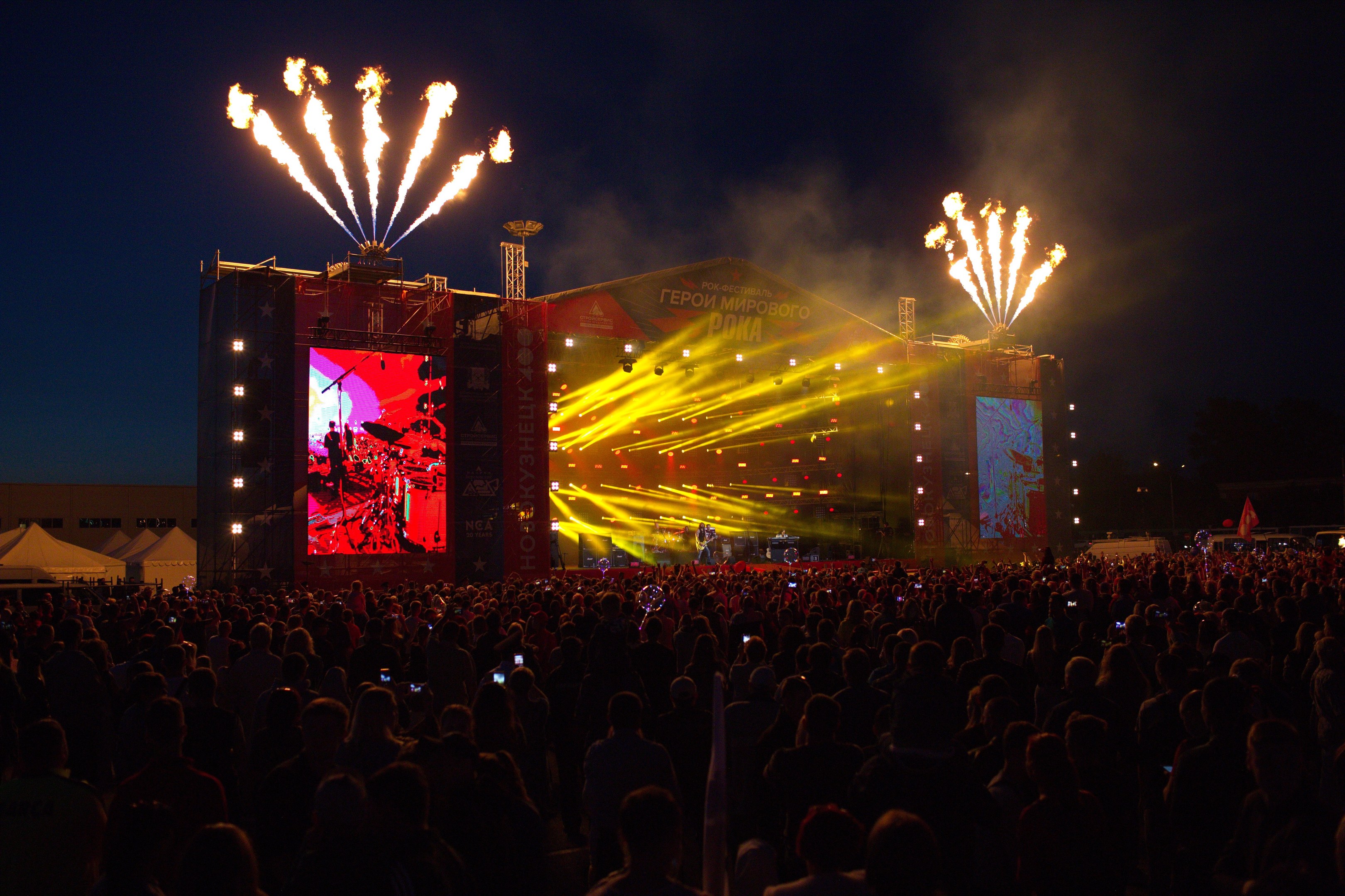 Eine große Menschenmenge auf einem Musikfestival vor einer hell erleuchteten Bühne, auf der Musiker spielen und von Bannern umgeben sind, mit Zelten links und Feuerwerk am dunklen Himmel dahinter.