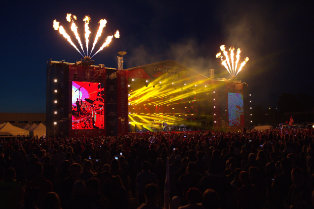 Eine große Menschenmenge auf einem Musikfestival vor einer hell erleuchteten Bühne, auf der Musiker spielen und von Bannern umgeben sind, mit Zelten links und Feuerwerk am dunklen Himmel dahinter.