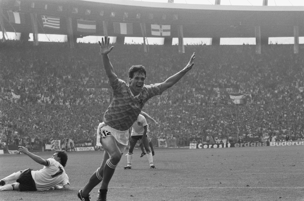 Ein Schwarz-Weiß-Foto eines Fussballspielers, der nach einem Tor feiert, umgeben von Menschen auf dem Boden, mit Brettern, Fahnen und Pfählen im Hintergrund unter einem klaren Himmel.