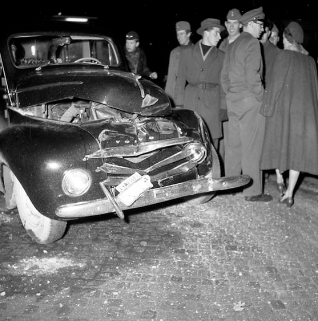 Ein Schwarz-Weiß-Bild eines verbeulten Autos am Straßenrand, umgeben von einer Gruppe besorgter Menschen.