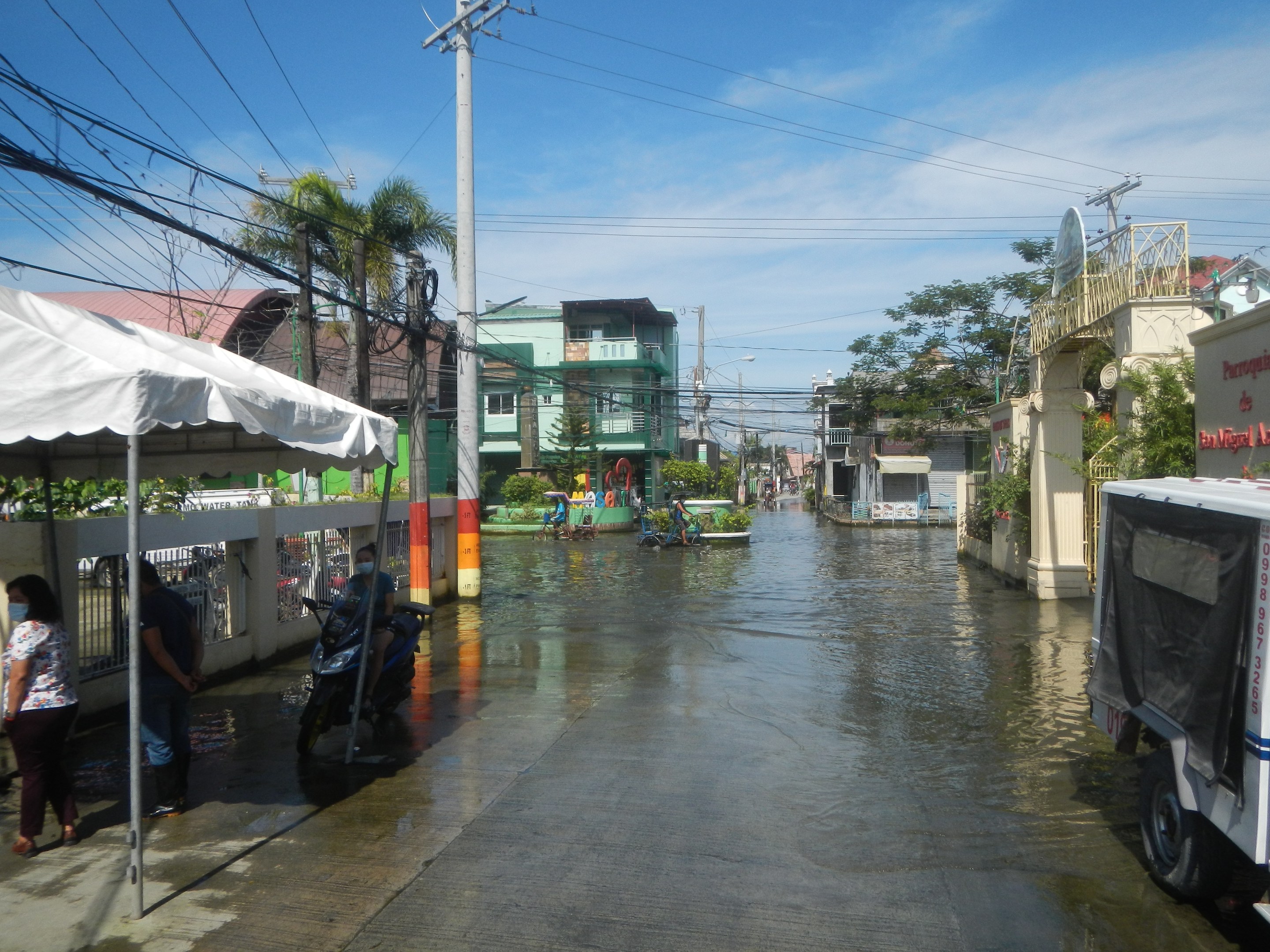 Überflutete Stadtstraße mit Fahrzeugen, Fußgängern, einem Zelt, Versorgungsmasten, Gebäuden, Bäumen und bewölktem Himmel.