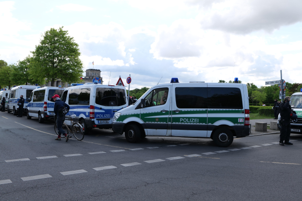 Eine Gruppe von Polizeiwagen, die an einer Straße geparkt sind, mit einem Radfahrer im Vordergrund und Gebäuden, Bäumen und Schildern im Hintergrund unter einem bewölkten Himmel.