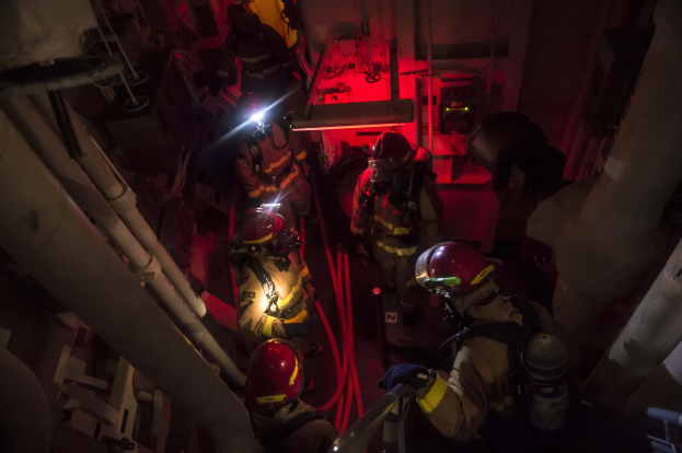 Feuerwehrleute in Helmen und Sauerstoffflaschen stehen in einem schwach beleuchteten Raum mit Rohren auf beiden Seiten und führen einen Rettungseinsatz in der Nähe von Maschinen und anderen Gegenständen durch.