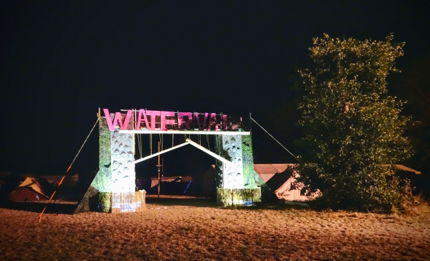 Bühne für ein nächtliches Musikfestival mit einem Tor mit Text, einem Baum rechts, Zelten im Hintergrund und einem dunklen Himmel.