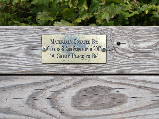 Holzbank mit einer Plakette, auf der "Materialien gespendet von Charles & Ann Schwabach 2017: Ein schöner Ort" steht, in einem Garten mit üppiger grüner Vegetation.