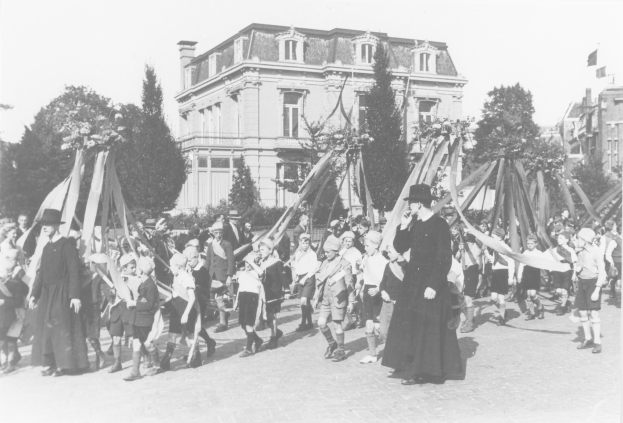 Schwarzes und weißes Bild einer Gruppe von Menschen, die eine Straße entlanggehen, einige halten Fahnen, tragen Hüte, es gibt Bäume, Gebäude und einen sichtbaren Himmel im Hintergrund, mit einem Maibaum, der auf ein Maifeierfest hinweist.