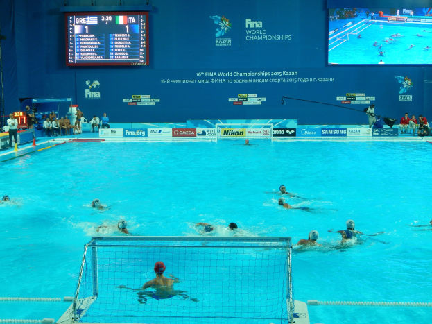 Eine Gruppe von Menschen, die Wasserball in einem großen Pool mit einem Netz spielen, Zuschauer auf der linken Seite und ein Bildschirm mit Text und Logos im Hintergrund bei den FINA-Weltmeisterschaften 2016 in Kazan.
