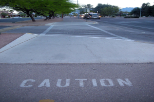 Eine Stadtstraße mit Fahrzeugen, ein Gehweg mit einem Warnschild neben einem Baum, Pfosten, Laternen, Bretter, Pflanzen, Gebäuden und Bergen unter einem klaren blauen Himmel.