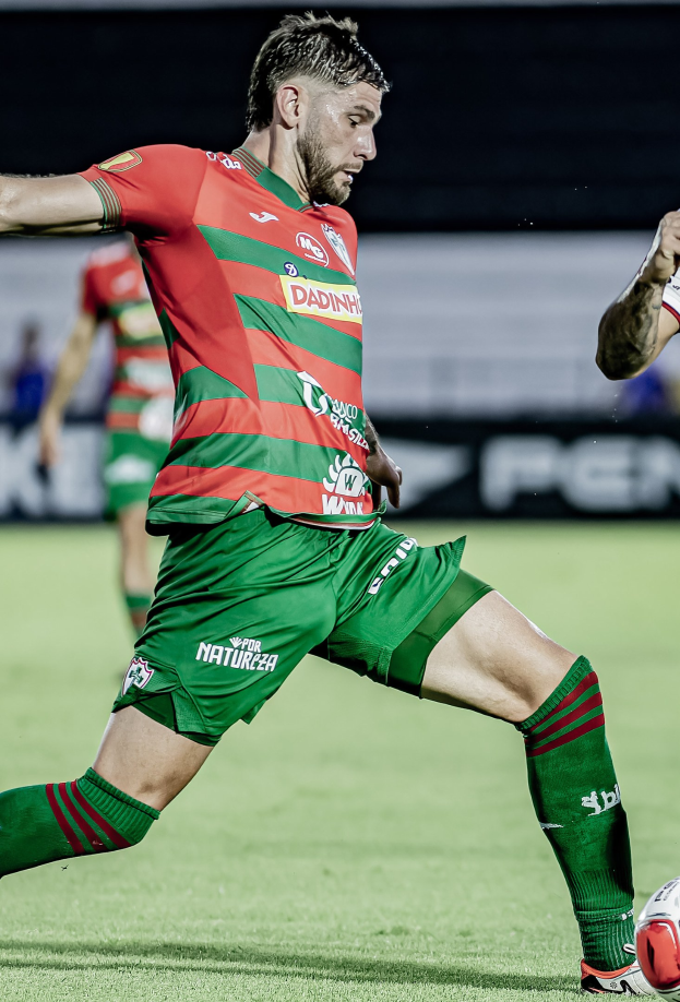 Ein Mann in roter und grüner Uniform kickt einen Fußball auf einem Rasenfeld, mit einer anderen Person im Hintergrund und einer Tafel mit der Aufschrift "Santos vs Vitoria Guimaraes."