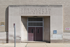 Das Südbildungshaus mit einer Tür, Schilder an der Wand und einem Weg, der zur Einfahrt führt.