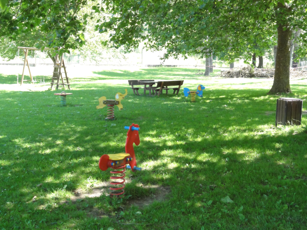 Ein Park mit Schaukeln, Bänken und einem Baum auf grünem Rasen, umgeben von grünem Wald.