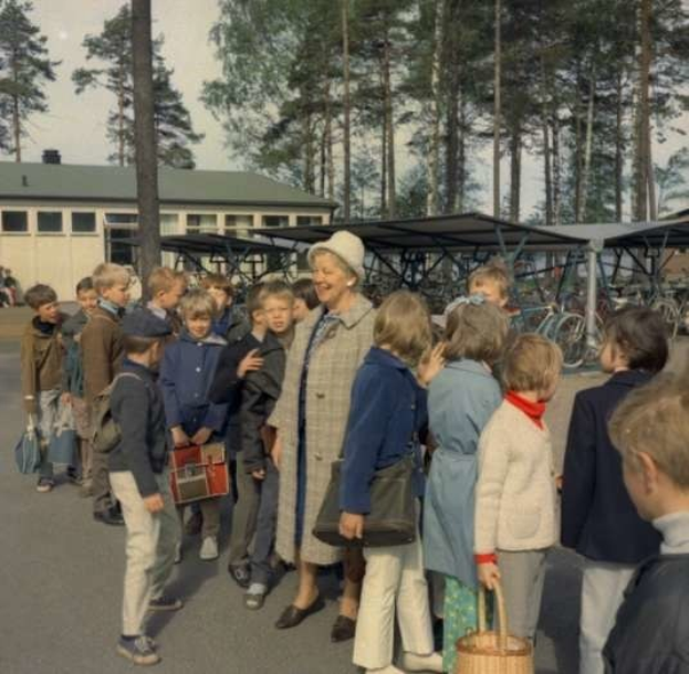 Eine Gruppe von Kindern steht vor einem Gebäude mit Fahrrädern und Schuppen im Hintergrund, unter einem klaren blauen Himmel.
