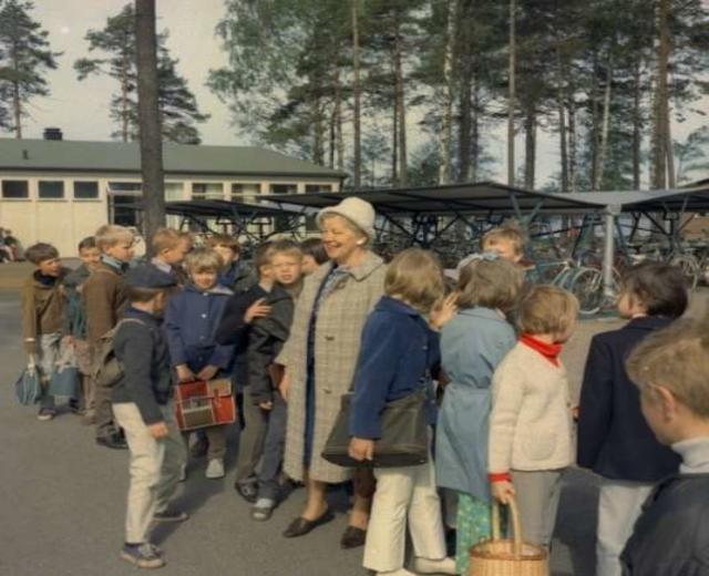 Eine Gruppe von Kindern steht vor einem Gebäude mit Fahrrädern und Schuppen im Hintergrund, unter einem klaren blauen Himmel.