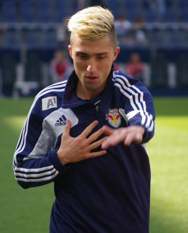 Ein junger Mann in einem blauen T-Shirt steht auf einem Fußballfeld und streckt die Arme aus, mit entschlossener Miene als Flügelspieler vor einem unscharfen Hintergrund.