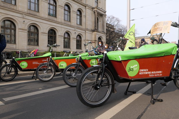 Eine Gruppe von Fahrrädern, die entlang einer Straße geparkt sind, mit einer Person in der Nähe und einem Fahrrad-Sharing-Banner im Vordergrund sowie Gebäuden, Bäumen und einem klaren Himmel im Hintergrund.