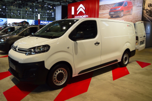 Ein weißer Citroën Berlingo Van wird auf einer Automesse gezeigt, umgeben von schwarzen Schlafzimmermöbeln, während die Wände mit Brettern und Lichtern geschmückt sind.