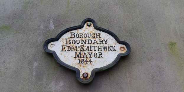 Metallplatte an der Außenseite eines Gebäudes, die die Borough-Grenze markiert, mit "Borough Boundary Edm Smithwick Mayor" in fetten weißen Buchstaben auf schwarzem Hintergrund.