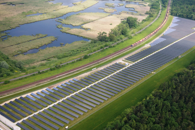 Luftaufnahme eines Solarparks mit Solarpanelen auf einem Feld, umgeben von Bäumen, Gras, Pflanzen und Wasser, mit einem Zug auf einer nahen Bahnlinie.