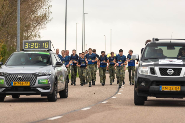 Gruppe von Menschen, die auf einer Straße neben parkenden Autos laufen, mit Bäumen auf der linken Seite und Strommasten im Hintergrund, unter einem sichtbaren Himmel, bei einem Europäischen Cross-Country-Challenge.