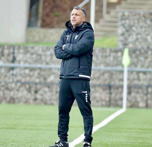 Ein Mann auf einem Fußballfeld mit verschränkten Armen, der eine schwarze Jacke und Hose trägt, vor einem Zaun, Treppe und Wand während eines Trainings.