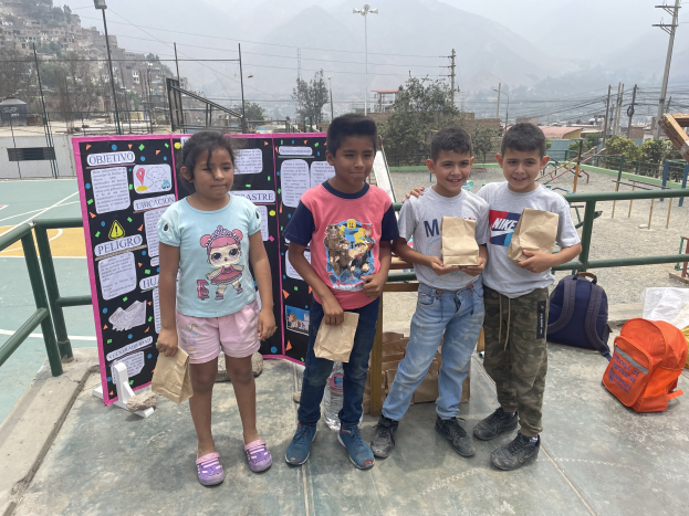 Gruppe von Kindern auf einem Basketballfeld mit Papierbeuteln in den Händen, mit Beuteln und Gegenständen rechts und einer Tafel mit Text links, vor dem Hintergrund von Zäunen, Pfählen, Straßenlaternen, Bäumen, Gebäuden, Hügeln und Himmel.