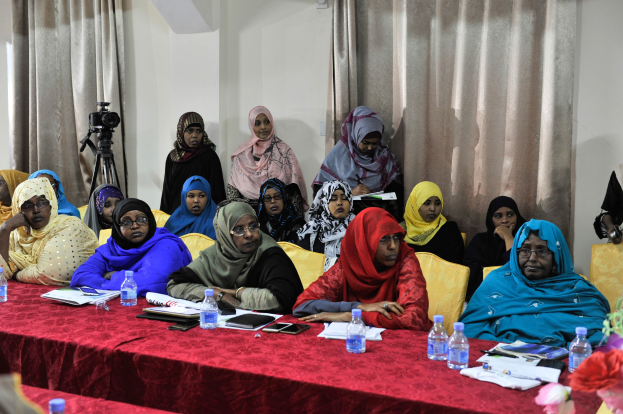 Eine Gruppe von Frauen, die an einem Tisch sitzen und verschiedene Gegenstände haben, diskutieren, mit einer Vase rechts und Vorhängen im Hintergrund.